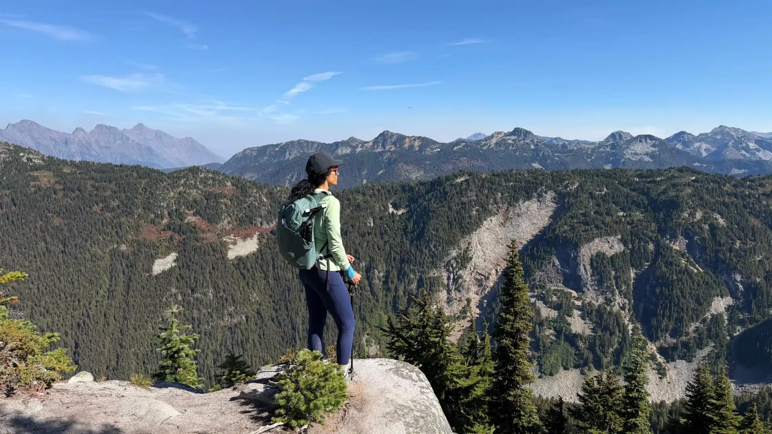 Hiker at a rocky viewpoint below Flora Peak’s summit overlooking forested slopes, scree and rugged mountain ridges
