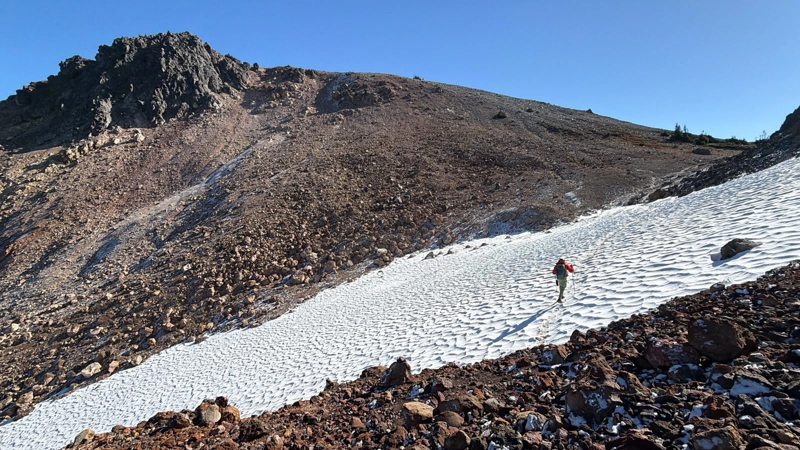 Hiker crossing a remnant snowfield beneath the rocky slopes approaching the Mount Price and Clinker col