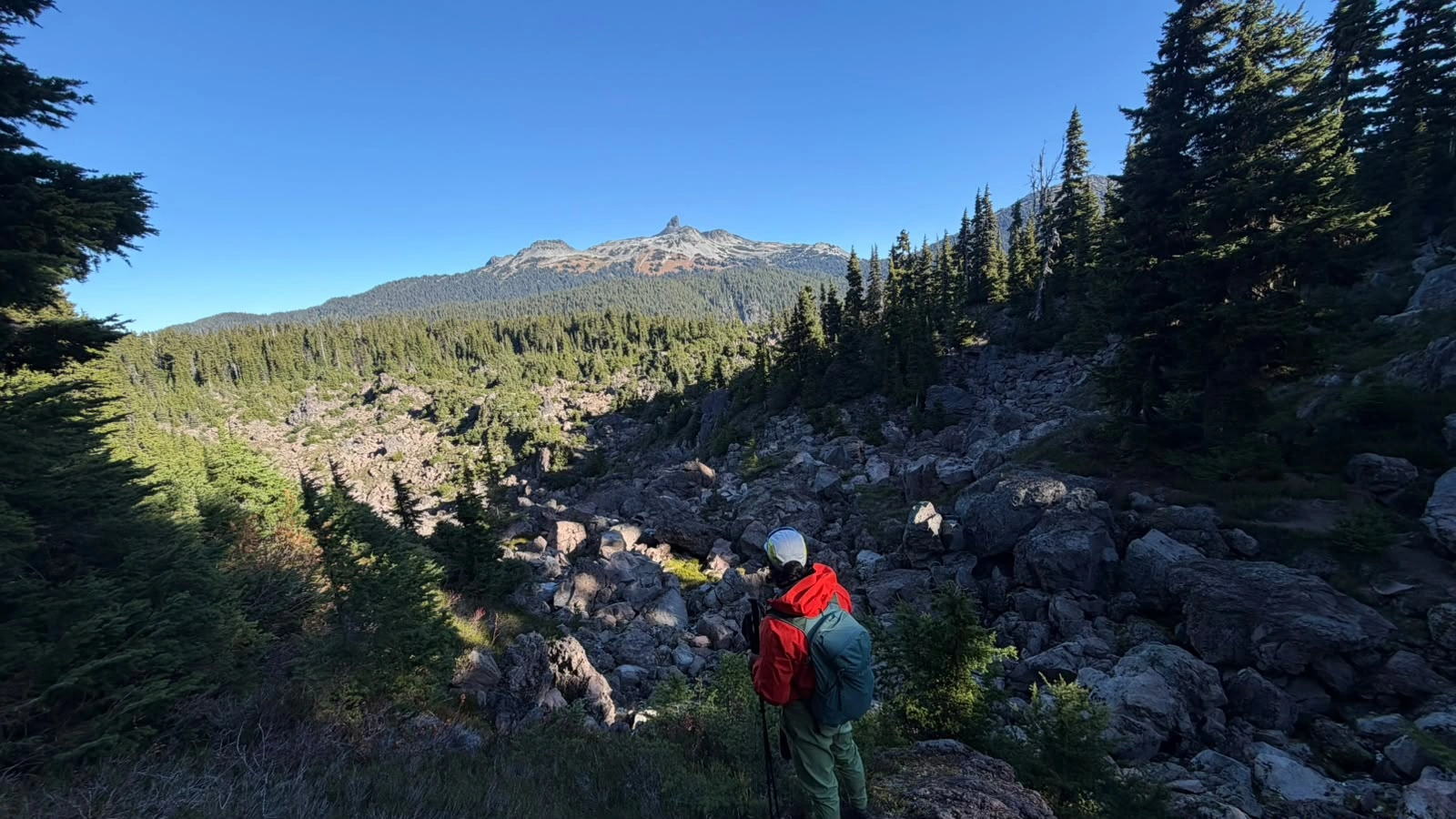 Hiker navigating a forested boulder field on the Mount Price approach, with Black Tusk in the distance